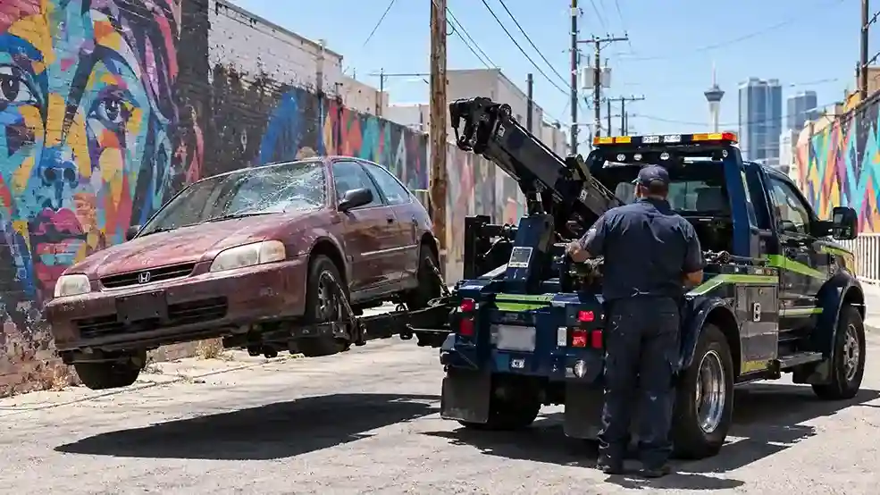 A wheel-lift tow truck lifting an old maroon hatchback in a downtown Las Vegas alley