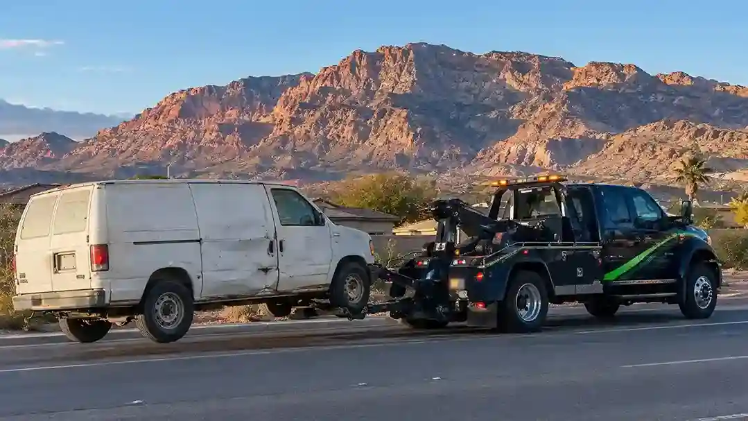 A wheel-lift tow truck towing an old white work van past Frenchman Mountain